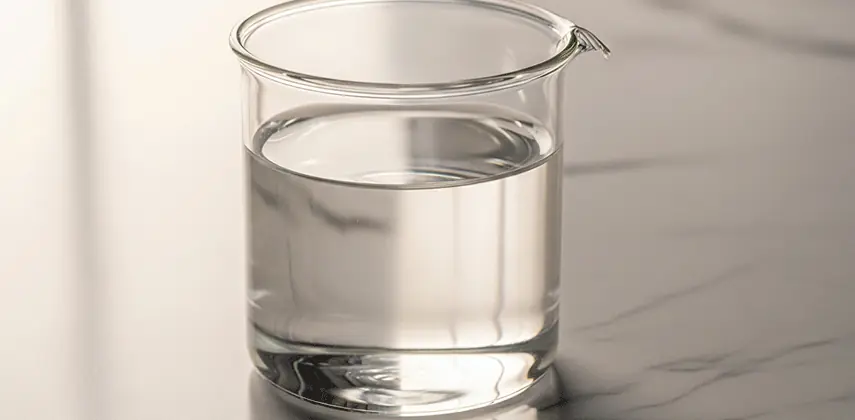 Şeffaf, viskoz bir sıvıyla (muhtemelen sıvı parafin) dolu, temiz bir laboratuvar cam kabı (beher). Minimalist beyaz mermer bir yüzey üzerinde, yumuşak stüdyo aydınlatması altında çekilmiş, ürün fotoğrafçılığı tarzında yüksek detaylı bir görsel